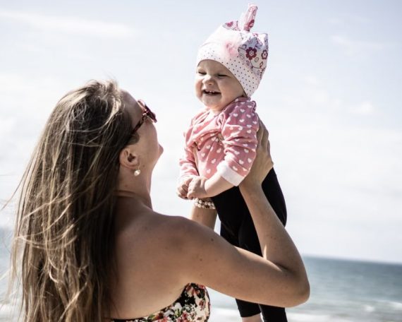 A happy mother lifting her smiling baby against a scenic beach backdrop.